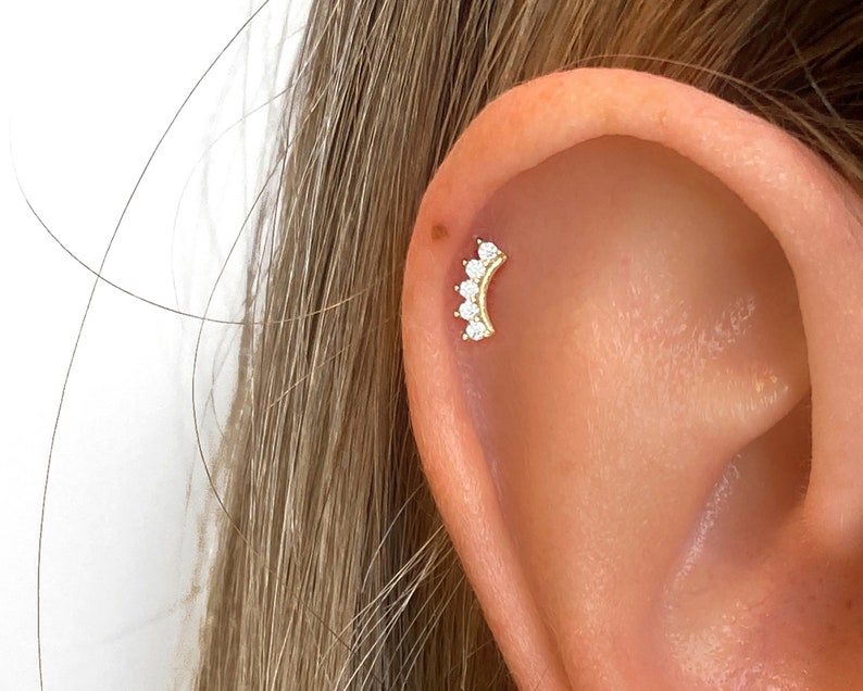 May include: A gold ear piercing with a crescent moon design featuring five small clear cubic zirconia stones.