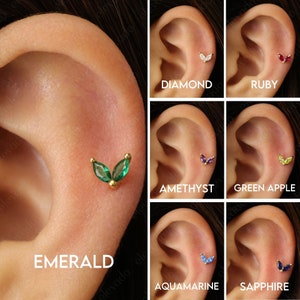 Może przedstawiać: Zbliżenie ucha z r&oacute;żnymi małymi, złotymi kolczykami w kształcie liści z r&oacute;żnokolorowymi kamieniami szlachetnymi. Kamienie szlachetne to szmaragd, diament, rubin, ametyst, zielone jabłko, akwamaryn i szafir.