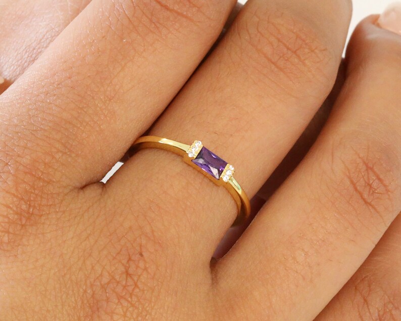 May include: A gold ring with a purple rectangular gemstone and two smaller clear gemstones on either side.