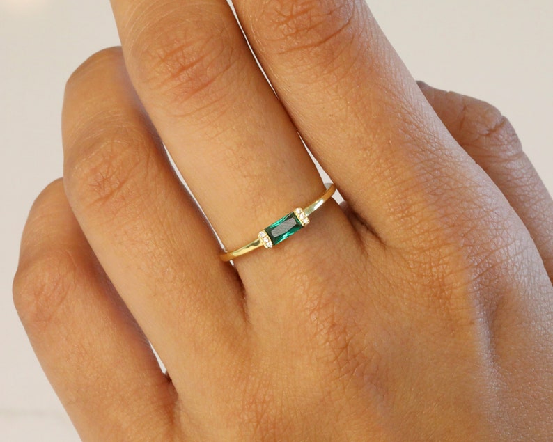 May include: A gold ring with a single emerald green rectangular stone set in the center. The ring is thin and delicate.