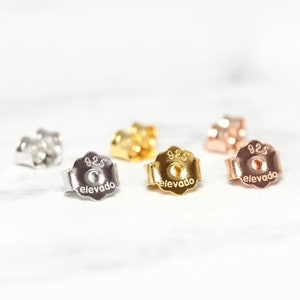 May include: Four earring backs in different colors: silver, gold, rose gold, and a darker gold. Each back is stamped with "925" and "elevado".