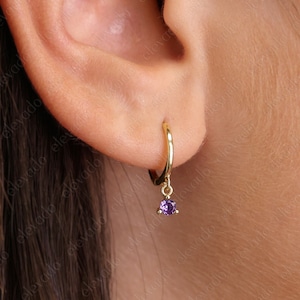K&ouml;nnte beinhalten: Goldene Creolen mit einem baumelnden violetten Edelstein. Die Ohrringe sind an einem Ohr einer Person zu sehen.