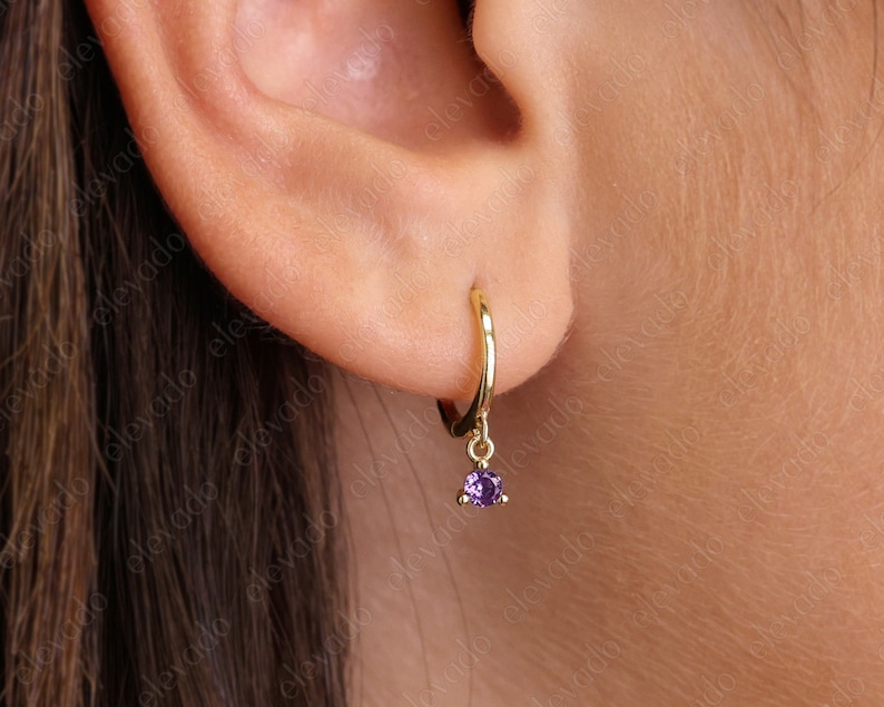 K&ouml;nnte beinhalten: Goldene Creolen mit einem baumelnden violetten Edelstein. Die Ohrringe sind an einem Ohr einer Person zu sehen.