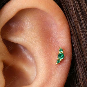 Op de afbeelding: Een close-up van een oor met een kleine gouden oorring met drie groene edelstenen. De oorring heeft de vorm van een halve maan.
