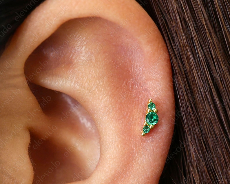 Op de afbeelding: Een close-up van een oor met een kleine gouden oorring met drie groene edelstenen. De oorring heeft de vorm van een halve maan.