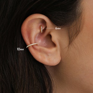 May include: Close-up of an ear adorned with two pieces of gold jewelry. A small hoop earring, 6mm in diameter, is in the upper ear. A 10mm ear cuff with a row of small, clear stones is on the outer ear. The jewelry sparkles.