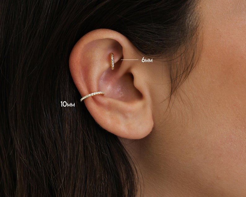 May include: Close-up of an ear adorned with two pieces of gold jewelry. A small hoop earring, 6mm in diameter, is in the upper ear. A 10mm ear cuff with a row of small, clear stones is on the outer ear. The jewelry sparkles.