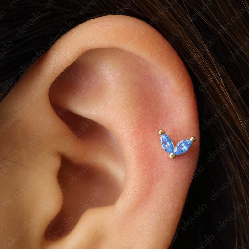 Może przedstawiać: Zbliżenie na ucho z małym złotym kolczykiem z dwoma niebieskimi kamieniami szlifowanymi w kształcie markizy.
