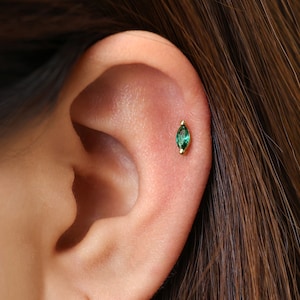 May include: A gold cartilage earring featuring a marquise-cut green gemstone set in a metal bezel. The earring is positioned on the upper ear cartilage, showcasing a slender, pointed oval shape with a vibrant emerald-toned stone.