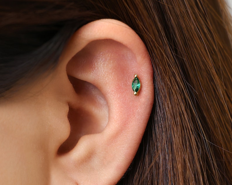 May include: A gold cartilage earring featuring a marquise-cut green gemstone set in a metal bezel. The earring is positioned on the upper ear cartilage, showcasing a slender, pointed oval shape with a vibrant emerald-toned stone.