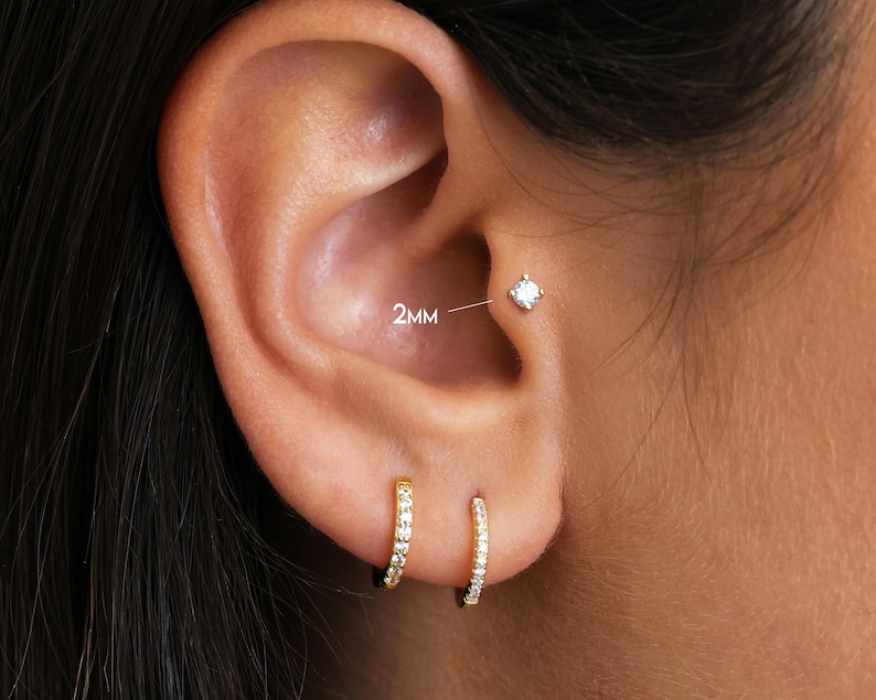 May include: Close-up of a person's ear with two gold hoop earrings with small clear stones. The top piercing has a single clear stone with the text "2MM" next to it.