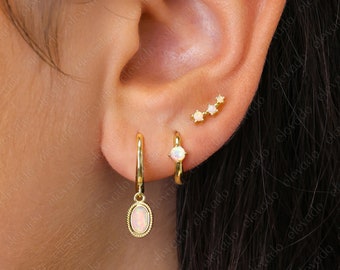 Könnte beinhalten: Goldene Creolen mit einem baumelnden ovalen Opal-Edelstein und einem kleineren Creolen-Ohrring mit einem einzelnen Opal-Edelstein. Eine kleine Reihe von drei Opal-Edelsteinen ist auch im oberen Ohr sichtbar.