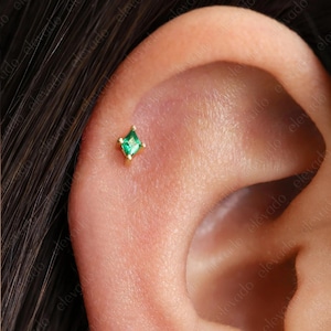 May include: A small, gold-colored stud earring with a square, emerald-green gemstone. The earring is set in a four-prong setting and is displayed on an ear. The earring is a delicate piece of jewelry.