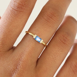 May include: A gold ring with a light blue rectangular gemstone and small clear gemstones on either side.