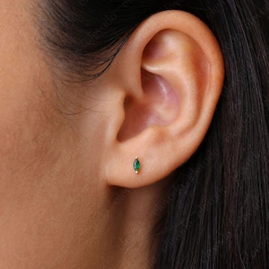 May include: A close-up of a person's ear with a small green gemstone earring. The earring is a simple stud with a small, oval-shaped green gemstone.