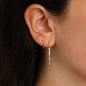 May include: A close-up of a person's ear with three gold earrings. The earrings are a small stud with a purple stone, a small hoop with purple stones, and a dangling sword charm with purple stones.