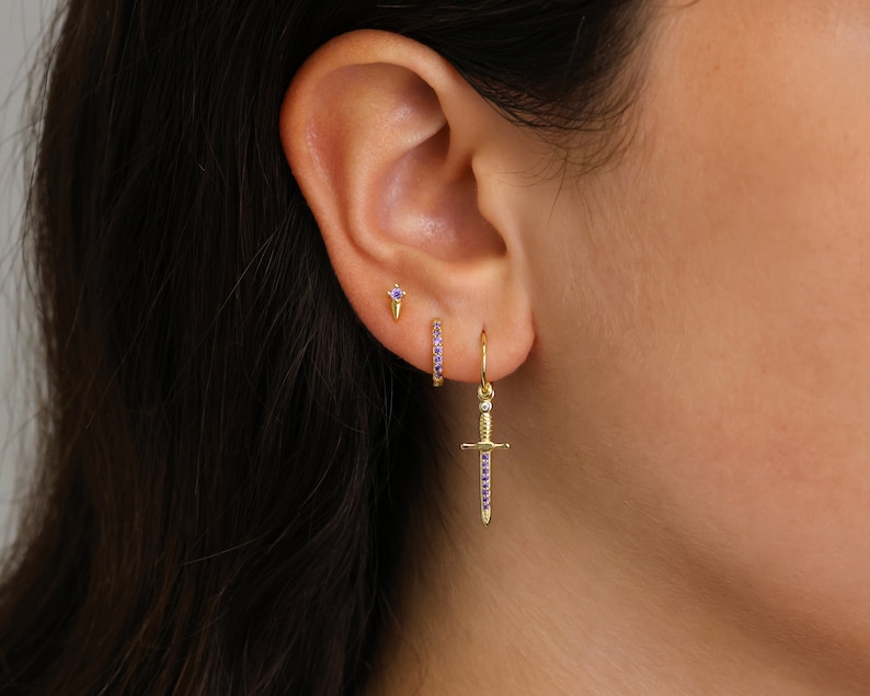 May include: A close-up of a person's ear with three gold earrings. The earrings are a small stud with a purple stone, a small hoop with purple stones, and a dangling sword charm with purple stones.