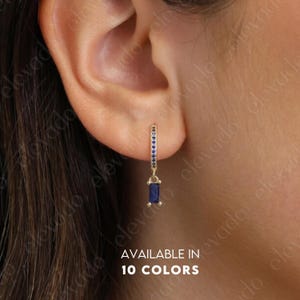 Może przedstawiać: Złoty kolczyk w kształcie koła z prostokątnym, ciemnoniebieskim wisiorkiem z kamienia szlachetnego. Koło ozdobione jest małymi niebieskimi kamieniami. Tekst "AVAILABLE IN 10 COLORS" jest widoczny na dole obrazu.