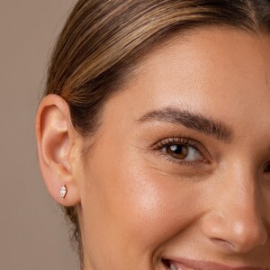 May include: A small gold stud earring featuring a marquise-cut clear gemstone. The earring is positioned in the earlobe, showcasing a slender, elongated shape with a pointed tip at both ends, secured by a thin gold metal setting.