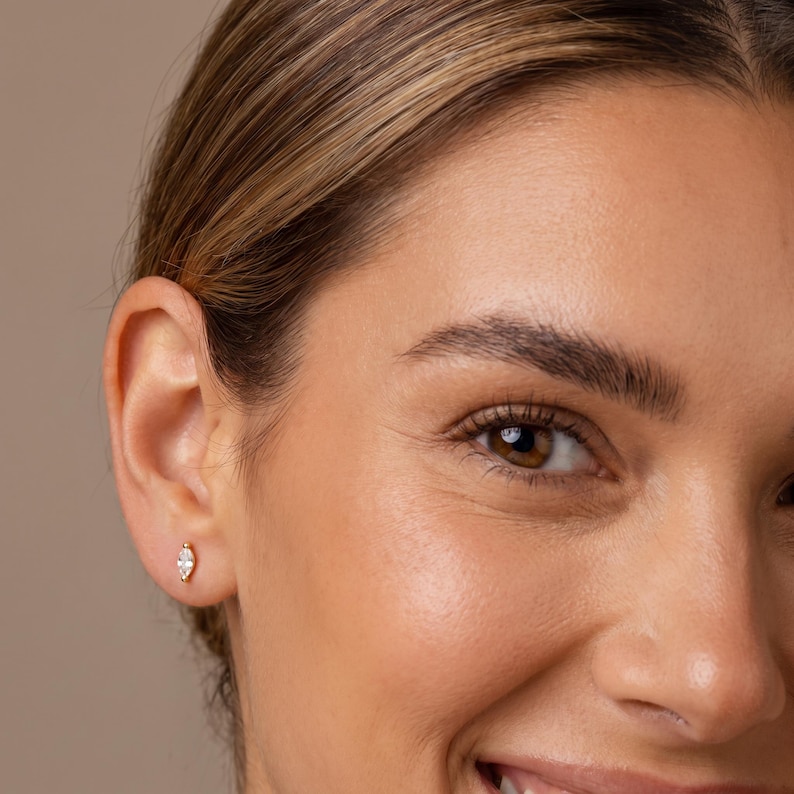 May include: A small gold stud earring featuring a marquise-cut clear gemstone. The earring is positioned in the earlobe, showcasing a slender, elongated shape with a pointed tip at both ends, secured by a thin gold metal setting.