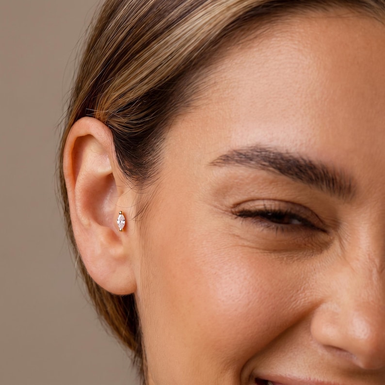 May include: A gold ear piercing stud featuring a small, marquise-cut clear gemstone. The jewelry is positioned in the conch area of the ear, showcasing a metallic gold setting that holds the elongated, pointed stone securely in place.