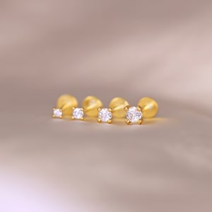 May include: Four gold-toned stud earrings with clear round gemstones. The earrings are arranged in a row on a light brown surface.