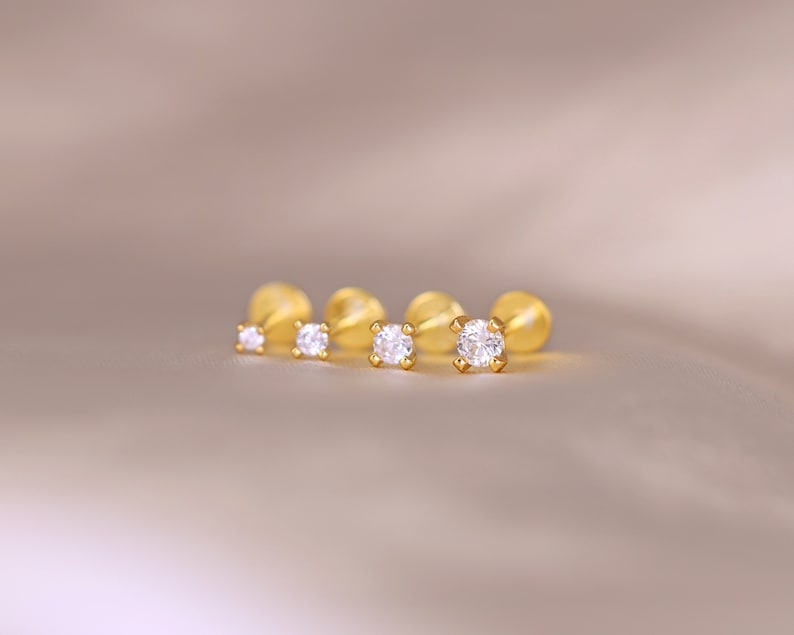 May include: Four gold-toned stud earrings with clear round gemstones. The earrings are arranged in a row on a light brown surface.