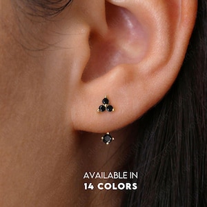 Puede incluir: Un par de pequeños pendientes con piedras preciosas negras. Los pendientes tienen forma de triángulo con tres piedras preciosas. Los pendientes están disponibles en 14 colores.