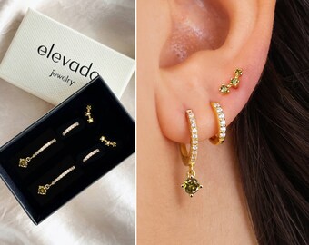 May include: A set of gold earrings with green gemstones and clear cubic zirconia stones. The earrings are in a black box with the word "elevada jewelry" printed on the lid.