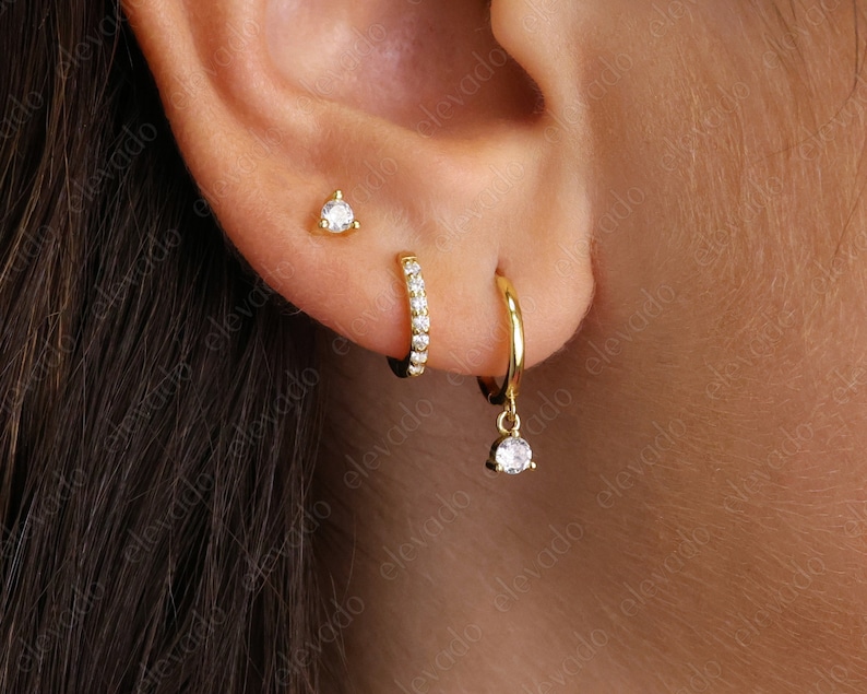 K&ouml;nnte beinhalten: Drei goldene Ohrringe mit kleinen, klaren Zirkonia-Steinen. Die Ohrringe sind ein einzelner Stein-Stecker, ein kleiner Reifen mit Steinen am Rand und ein gr&ouml;&szlig;erer Reifen mit einem baumelnden Stein.