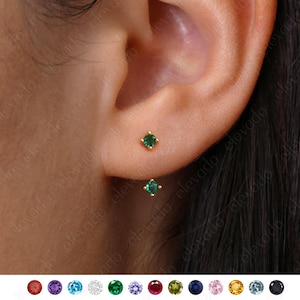 Peut inclure: Une paire de petites boucles d'oreilles en or avec des accents de pierres précieuses vertes. Les boucles d'oreilles sont présentées dans une image rapprochée de l'oreille d'une personne.