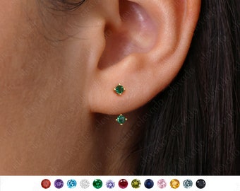 May include: A pair of small, gold stud earrings with green gemstone accents. The earrings are shown in a close-up image of a person's ear.