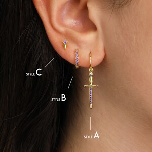 May include: Three different styles of earrings with purple gemstones. Style A is a dangling sword earring, Style B is a small hoop earring, and Style C is a small stud earring.
