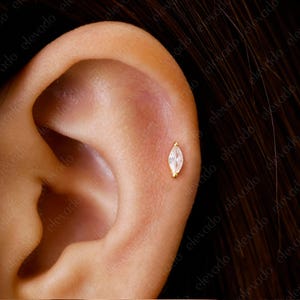 以下が含まれることがあります： 金色の枠にセットされた、小さな透明なマルキーズ型の宝石のスタッドピアスをした人の耳のクローズアップ。