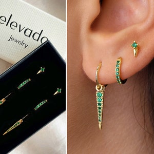 May include: A set of gold earrings with green gemstones. The earrings include a pair of small studs, a pair of small hoops, and a pair of dangle earrings with a pointed design. The earrings are displayed in a black box with the text "elevada jewelry".