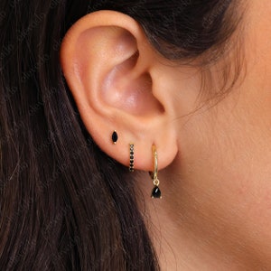 May include: A set of three gold earrings worn in an earlobe. The first is a small marquise-cut black stone stud, the second is a gold hoop encrusted with tiny black stones, and the third is a gold hoop featuring a dangling pear-cut black stone.