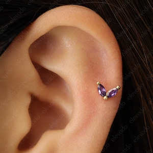 Może przedstawiać: Zbliżenie ucha z małym złotym kolczykiem z dwoma fioletowymi kamieniami szlachetnymi w kształcie litery V.