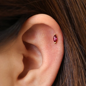 May include: A gold cartilage earring featuring a marquise-cut red gemstone set vertically. The jewelry has a small gold bead at both the top and bottom ends of the stone, securing it in place against the upper ear cartilage.