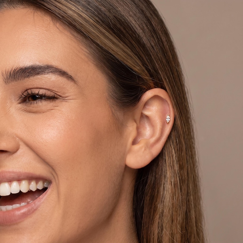 May include: A small, marquise-cut clear gemstone stud earring set in a metallic post, worn in the upper cartilage of the ear. The earring features a pointed, elongated shape with a single faceted stone that reflects light, secured by a metal backing.