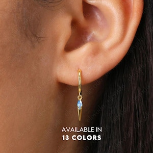 Puede incluir: Aretes de aro de oro con una pequeña piedra preciosa azul que cuelga de la parte inferior. Los aretes están disponibles en 13 colores. El texto "AVAILABLE IN 13 COLORS" es visible debajo de los aretes.