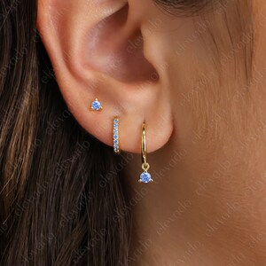 K&ouml;nnte beinhalten: Drei goldene Ohrringe mit blauen Edelsteinen. Die Ohrringe sind ein kleiner Stecker, ein kleiner Ring mit blauen Edelsteinen und ein Ring mit einem blauen Edelstein, der herunterh&auml;ngt.