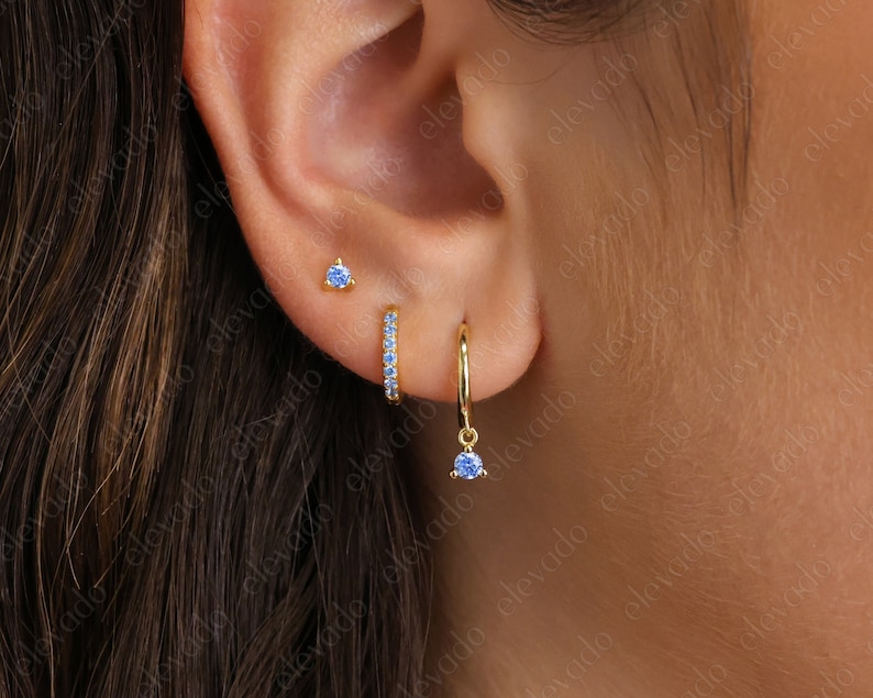 K&ouml;nnte beinhalten: Drei goldene Ohrringe mit blauen Edelsteinen. Die Ohrringe sind ein kleiner Stecker, ein kleiner Ring mit blauen Edelsteinen und ein Ring mit einem blauen Edelstein, der herunterh&auml;ngt.