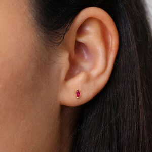 May include: A close-up of a person's ear with a small, red gemstone stud earring. The earring is set in gold and has a marquise shape.