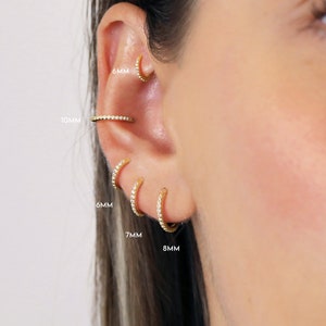 May include: A close-up of a person's ear with five different sized gold hoop earrings with small clear stones. The earrings are labeled with their sizes in millimeters: 6mm, 7mm, 8mm, 10mm, and 6mm.
