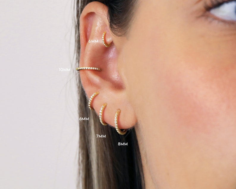May include: A close-up of a person's ear with five different sized gold hoop earrings with small clear stones. The earrings are labeled with their sizes in millimeters: 6mm, 7mm, 8mm, 10mm, and 6mm.