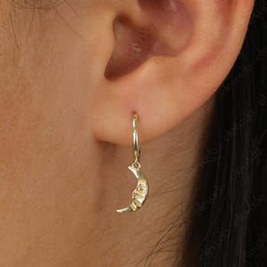 May include: Gold hoop earrings with a crescent moon charm. The moon charm has a face on it.