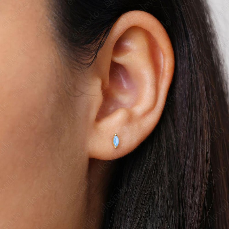 May include: A close-up of a person's ear with a small, light blue marquise-shaped opal stud earring in a gold setting.