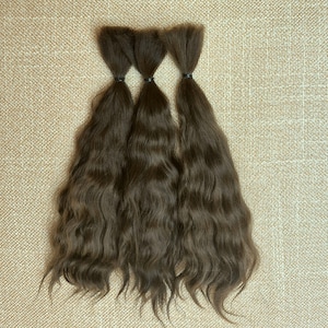 Puede incluir: Tres paquetes de extensiones de cabello castaño medio, cada uno asegurado en la parte superior con una pequeña banda elástica negra. El cabello tiene una textura ondulada natural y cae en mechones largos y sueltos, mostrando un color castaño uniforme.