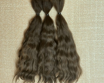 May include: Three bundles of medium brown hair extensions, each secured at the top with a small black elastic band. The hair has a natural wavy texture and flows downward in long, loose strands, displaying a consistent, uniform brown color throughout.