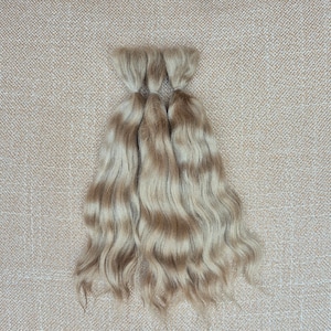 Puede incluir: Tres paquetes de extensiones de cabello ondulado en tono rubio ceniza claro. Cada paquete está asegurado en la parte superior con una banda, presentando una mezcla multitonal de reflejos rubios fríos y castaños claros con dimensión.